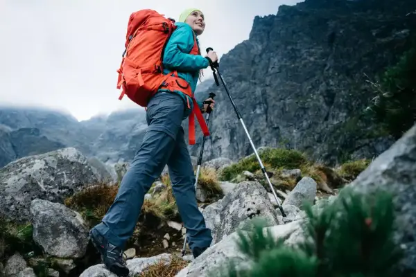 Kobieta z plecakiem wyrusza na wypad w góry, wspierając się kijkami trekkingowymi po kamienistym szlaku.