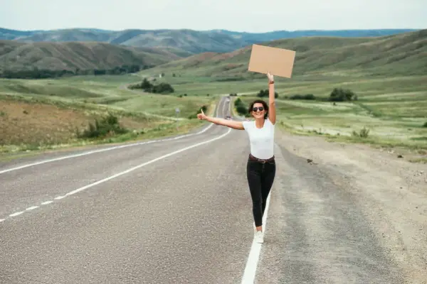 Kobieta z kartonem i kciukiem w górze na pustej drodze. Uczy się, jak łapać stopa, podróżując przez malownicze krajobrazy.